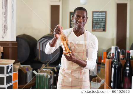 Smiling salesman in apron proposing wine in bottles in winery shop Smiling salesman in apron proposing wine in bottles in winery shop 66402633