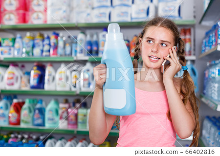 concentrated tween girl talking on phone and looking at bottle with household chemicals while in supermarket concentrated tween girl talking on phone and looking at bottle with household chemicals while in supermarket 66402816