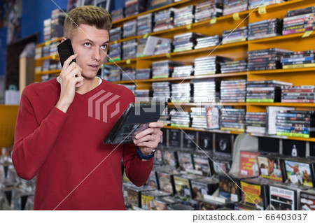 Guy customer talking by phone near shelves with DVDs in shop Guy customer talking by phone near shelves with DVDs in shop 66403037