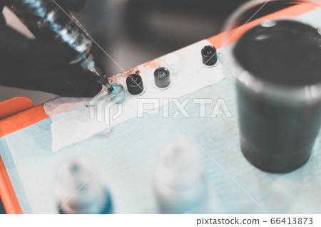 Tattoo salon. Tattoo machine and prepared paints for tattoos on the table. The process of curing paint. Close-up, tinted, tattooist. Tattoo salon. Tattoo machine and prepared paints for tattoos on the table. The process of curing paint. Close-up, tinted, tattooist. 66413873