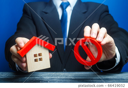 A man holds a house and the red prohibition symbol NO. Bank refuses to provide a mortgage loan. Housing problems. Confiscation pledged property. Prohibition of operation. Denial of insurance payment. A man holds a house and the red prohibition symbol NO. Bank refuses to provide a mortgage loan. Housing problems. Confiscation pledged property. Prohibition of operation. Denial of insurance payment. 66419032
