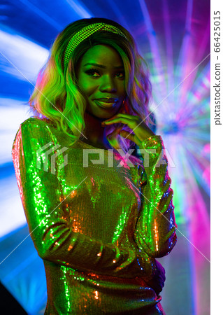 Pretty african woman seductively smiles touching face with her hand while looking away. Silhouette of stylish model in bright colors of unusual neon lighting. Vogue concept. Beauty concept Pretty african woman seductively smiles touching face with her hand while looking away. Silhouette of stylish model in bright colors of unusual neon lighting. Vogue concept. Beauty concept 66425015