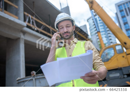 Building worker in helmet and yellow vest holding blueprint, talking on the phone 66425338