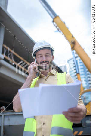 Building worker in helmet and yellow vest holding blueprint, talking on the phone, smiling 66425339