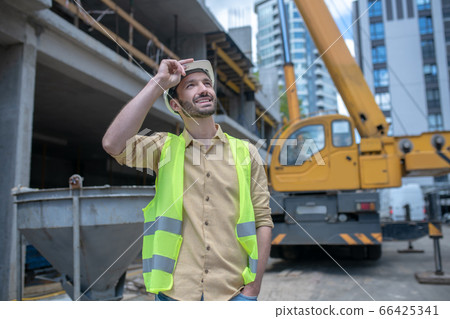 Building worker in helmet and yellow vest touching his helmet with his hand, looking up Building worker in helmet and yellow vest touching his helmet with his hand, looking up 66425341