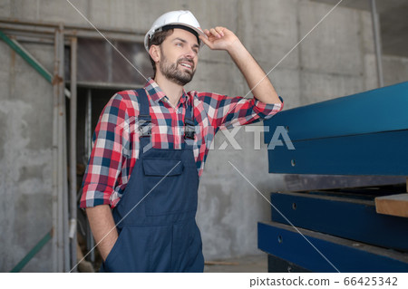 Building worker in helmet and checked shirt standing with one hand in pocket, touching his helmet with another 66425342