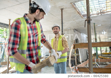 Building workers in yellow vests and gloves carrying together wooden board Building workers in yellow vests and gloves carrying together wooden board 66425380