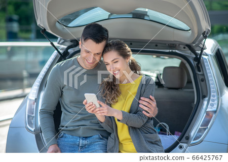 Woman with smartphone and man crouched on car trunk. 66425767