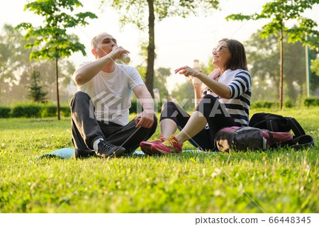 Happy mature couple sitting in the park on fitness mat, resting drinking yogurt after sports exercises. Happy mature couple sitting in the park on fitness mat, resting drinking yogurt after sports exercises. 66448345