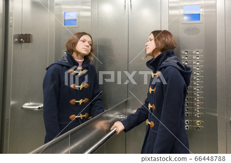 Teenage girl standing in elevator, looking in the mirror 66448788