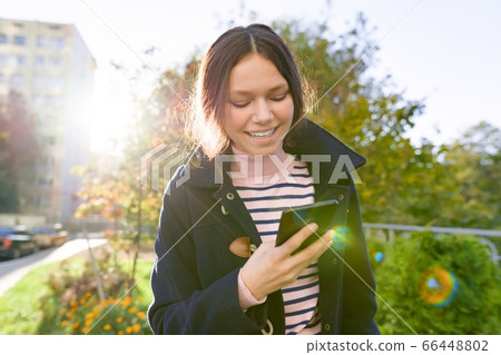 Teen girl in coat using smartphone, sunny autumn day Teen girl in coat using smartphone, sunny autumn day 66448802