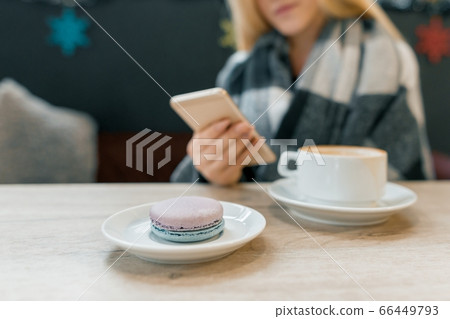 Macaroon in plate, cup of coffee, beautiful blonde girl in woolen blanket with smartphone 66449793
