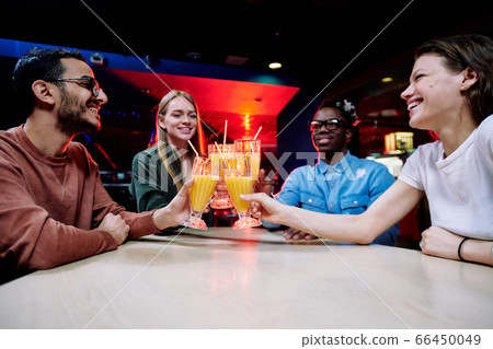 Happy young intercultural couple clinking with glasses of orange juice by table Happy young intercultural couple clinking with glasses of orange juice by table 66450049