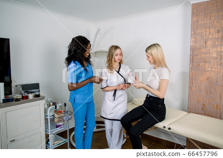 Consultation in cosmetology clinic. wo female professional beauty doctors, African and Caucasian women in medical suits, talking with pretty smiling Caucasian woman patient and showing new treatment Consultation in cosmetology clinic. wo female professional beauty doctors, African and Caucasian women in medical suits, talking with pretty smiling Caucasian woman patient and showing new treatment 66457796