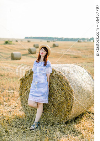 Countryside landscape of farm wheat field at summer sunset. Pretty young happy Caucasian woman in straw hat and blue striped dress posing near the hay bale and enjoying warm evening and vacations Countryside landscape of farm wheat field at summer sunset. Pretty young happy Caucasian woman in straw hat and blue striped dress posing near the hay bale and enjoying warm evening and vacations 66457797