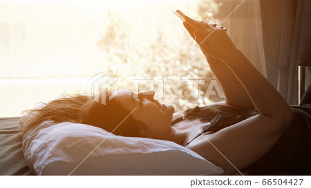 pretty woman chats on smartphone lying in bed bright window 66504427