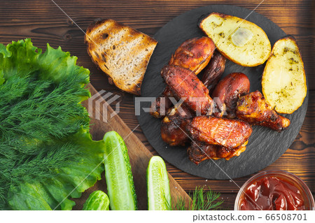 Grilled chicken drumsticks and wings on a slate dish with fresh cucumbers, ketchup, baked potatoes and herbs on a wooden table, top view, flat lay 66508701
