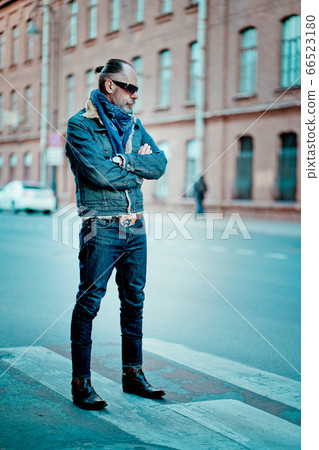 a man walks in the street, he has denim jacket, glasses, mustache and beard 66523180