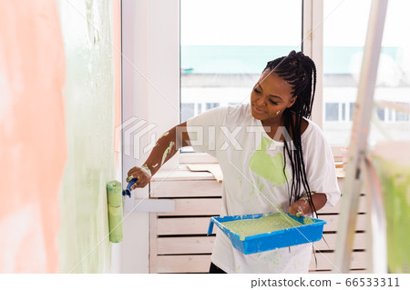 Happy African American woman painting wall in her new apartment. Renovation, redecoration and repair concept. Happy African American woman painting wall in her new apartment. Renovation, redecoration and repair concept. 66533311