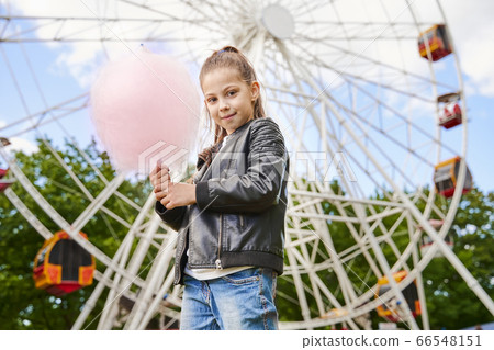 Portrait of a child with sweet cotton candy. A little girl on the background of ferris wheel is eating candy-floss summer sunny day. Portrait of a child with sweet cotton candy. A little girl on the background of ferris wheel is eating candy-floss summer sunny day. 66548151
