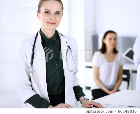 Doctor woman at work with patient and colleague at background. Physician filling up medical documents or prescription while standing in hospital reception desk. Data in medicine and health care 66549836