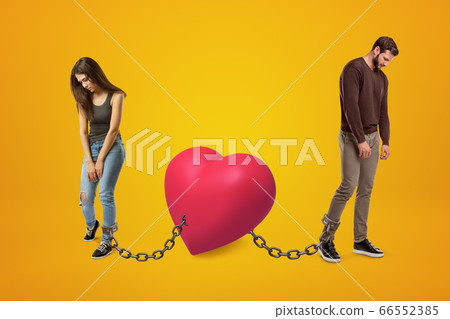 Young sad man and woman wearing casual clothes chained to pink heart on yellow background Young sad man and woman wearing casual clothes chained to pink heart on yellow background 66552385