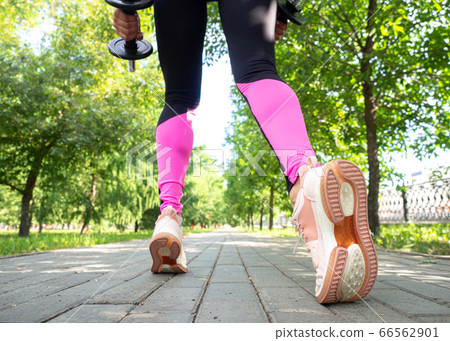 Female legs and feet in leggings and sneakers. Faceless woman does step forward with dumbbells in public park outdoor in summer, wide angle, back view, selective focus. 66562901
