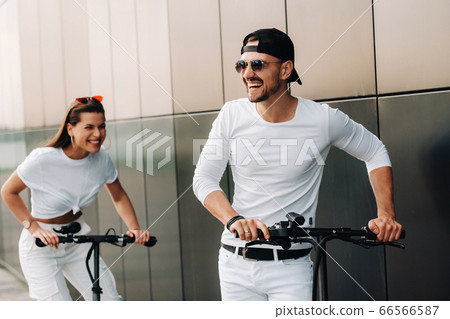 A girl and a guy are walking on electric scooters around the city, a couple in love on scooters. A girl and a guy are walking on electric scooters around the city, a couple in love on scooters. 66566587