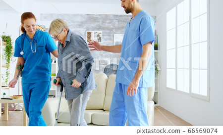 Female doctor and her assistant helping old woman with crutches to stand up from the couch Female doctor and her assistant helping old woman with crutches to stand up from the couch 66569074