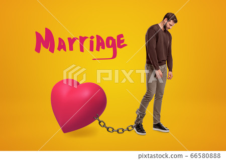 Young sad man wearing casual clothes chained to pink heart with 'Marriage' sign above on yellow background Young sad man wearing casual clothes chained to pink heart with 'Marriage' sign above on yellow background 66580888