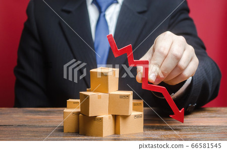 Businessman is holding a red arrow down above boxes stack pile. Business industrial production drop. Decrease freight transportation, volumes of delivery of products. Overproduction, scarcity of goods Businessman is holding a red arrow down above boxes stack pile. Business industrial production drop. Decrease freight transportation, volumes of delivery of products. Overproduction, scarcity of goods 66581545