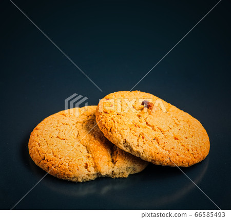 Two oatmeal cookies lie on top of each other and one of them is bitten. The photo is on a black background with reflections. 66585493