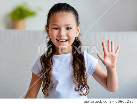 Cute Asian Kid Girl Waving Hand Sitting On Sofa Indoors Cute Asian Kid Girl Waving Hand Sitting On Sofa Indoors 66603285