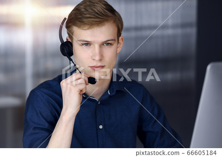 Young blond businessman using headset and computer in a darkened office, glare of light on the background. Startup business means working hard Young blond businessman using headset and computer in a darkened office, glare of light on the background. Startup business means working hard 66605184