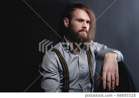 A portrait of an attractive brooding bearded man sitting against a wall looks nowhere and reflects on the dynocy and other problems. Studio portrait of stylish hipster 66605528