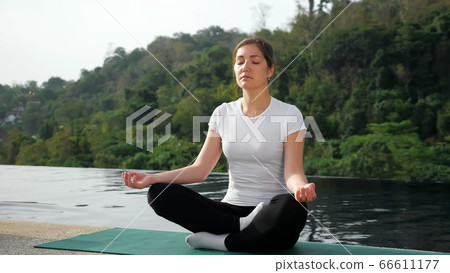 Novice female yogi sitting in lotus position, back to the pool 66611177