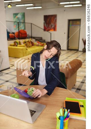 Girl talking on smartphone, behind laptop with fork and lunchbox. 66616246