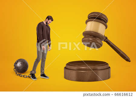 Distressed young man chained to shackle ball, looking down and walking slowly toward huge brown sound block with gavel on amber background. Distressed young man chained to shackle ball, looking down and walking slowly toward huge brown sound block with gavel on amber background. 66616612