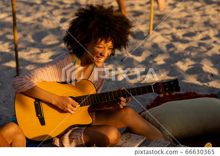 Young mixed race woman playing guitar and laughing on beach 66630365