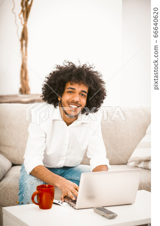 Arab student works laptop at coffee table with red cup on it. Handsome man smiles siiting at home sofa. Home education concept. Tinted image Arab student works laptop at coffee table with red cup on it. Handsome man smiles siiting at home sofa. Home education concept. Tinted image 66640620