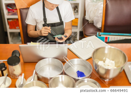 Female baker using cell phone with laptop and bakery ingredient on table. Female baker using cell phone with laptop and bakery ingredient on table. 66649491