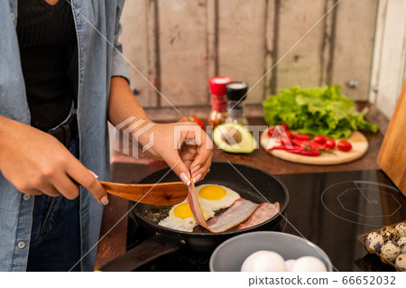 Young female standing by electric stove and putting slice of bacon on frying pan 66652032
