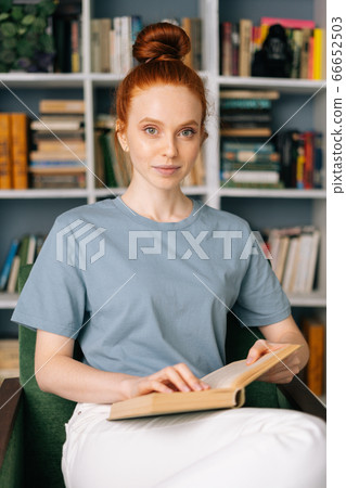 Loving redhead young woman student is reading book enjoys of rest at home office, looking at camera. 66652503