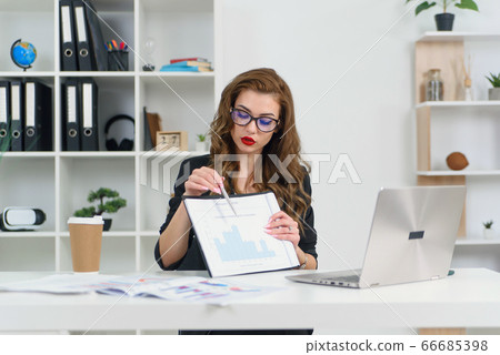 Experienced successful business lady in stylish clothes and glasses talking with her business partner by video call and shows report with graphs. 66685398