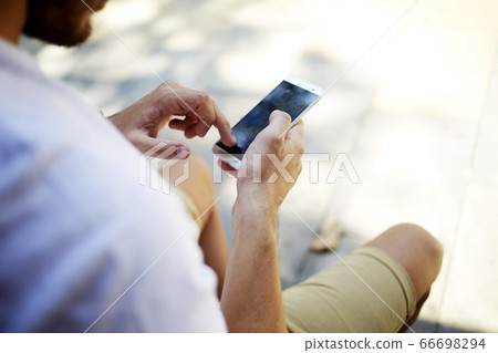 Hipster guy searching information on cell telephone while sitting outdoors Hipster guy searching information on cell telephone while sitting outdoors 66698294