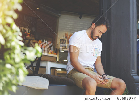Hipster guy searching information on cell telephone while sitting outdoors Hipster guy searching information on cell telephone while sitting outdoors 66698977