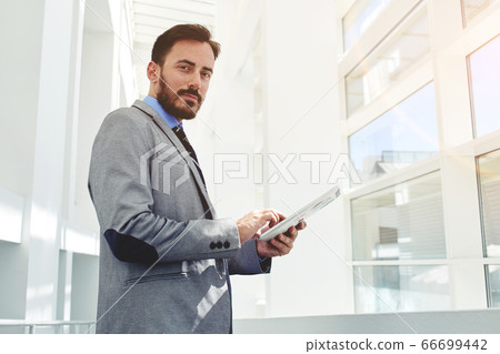Half length portrait of a young serious man bookkeeper holding touch pad while standing in modern interior, 66699442