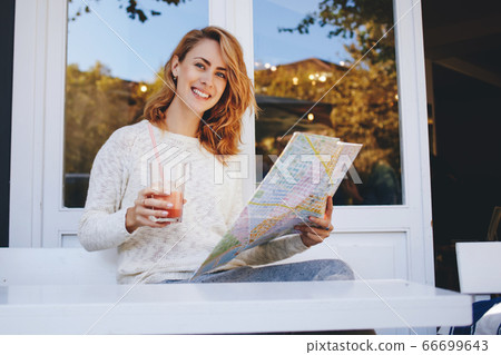 Young female tourist holding in hands map and glass of juice while sitting in modern sidewalk cafe, 66699643