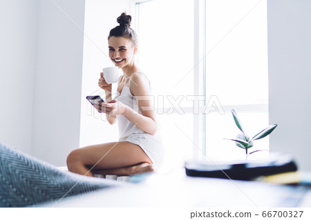 Cheerful woman drinking coffee and surfing smartphone at window sill at home 66700327