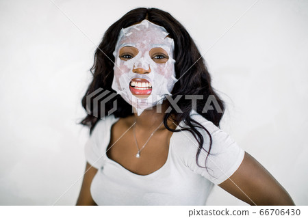 Happy smiling African young woman doing facial sheet mask, posing to camera on white background. Beauty and Skin Care Concept. Woman with cleansing mask on her face. Copy space 66706430
